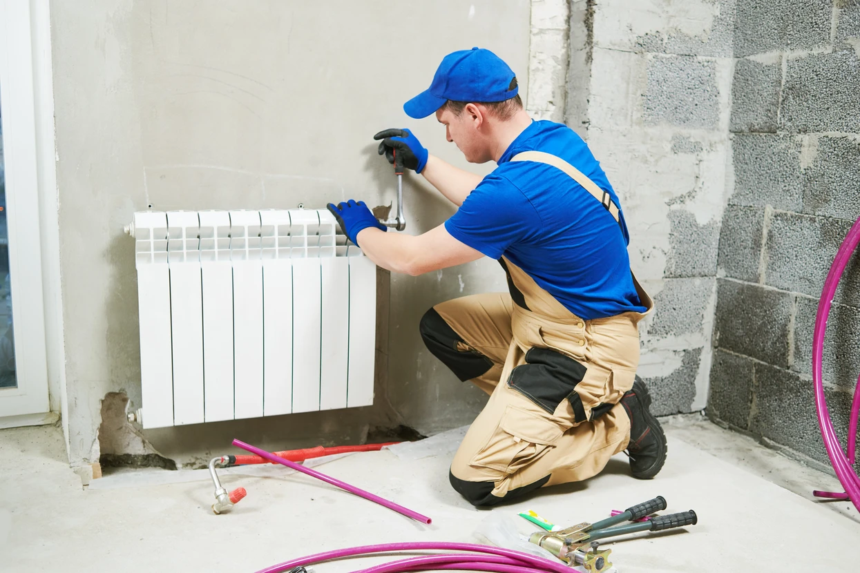 Plombier en uniforme bleu installant un radiateur à ailettes blanches dans une maison en construction